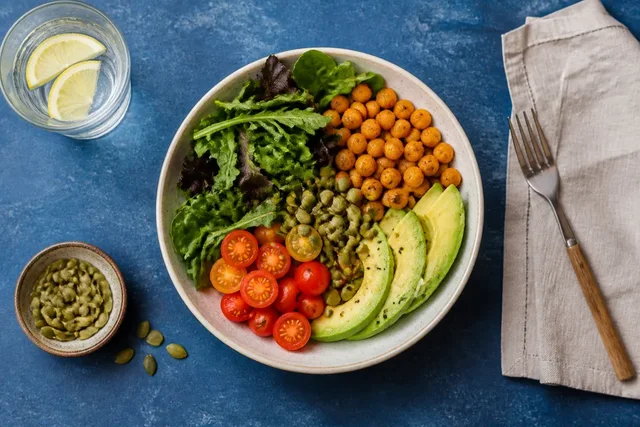 Illustrative overhead scene of a bowl with greens, chickpeas, tomatoes, avocado and seeds on a blue table.