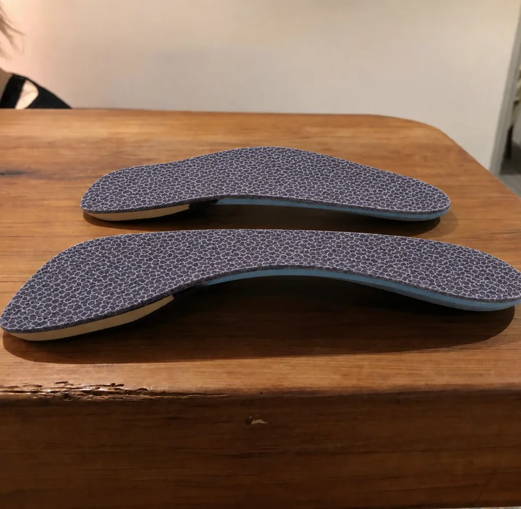 A pair of custom foot orthotics with charcoal-grey honeycomb-patterned covers on a wooden table.
