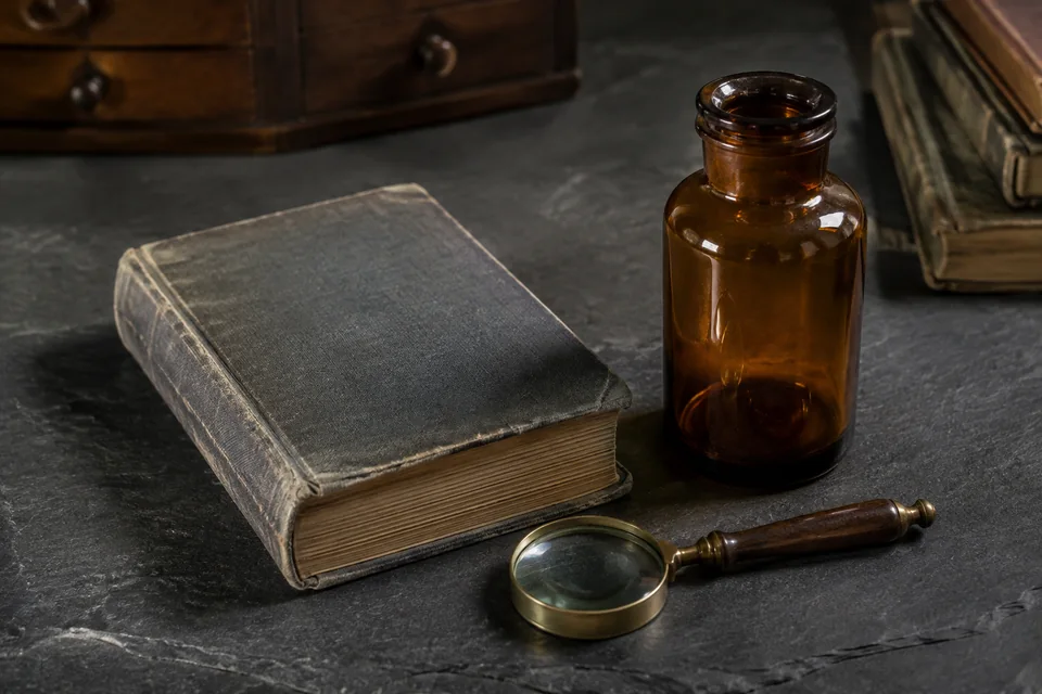 Illustrative still life of a vintage clothbound book, unlabelled brown glass bottle and magnifying glass.