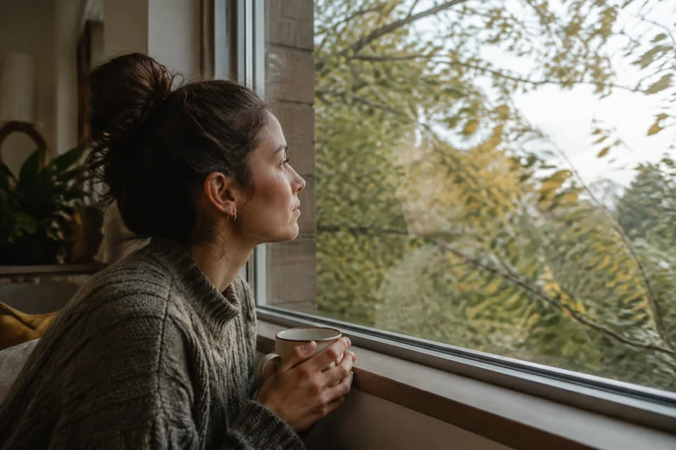 Illustrative photograph of a woman holding a mug and looking through a home window at leafy trees.