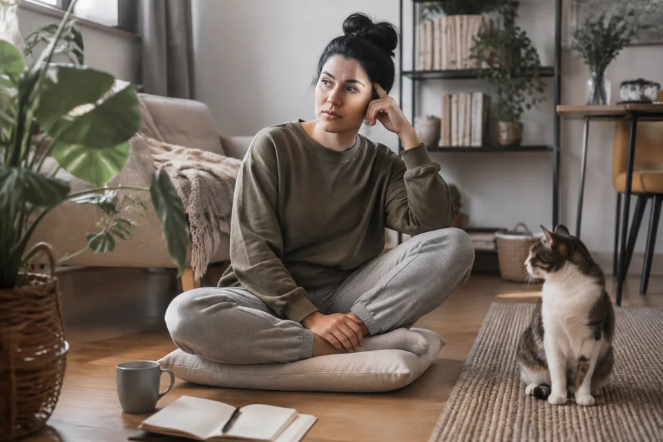 Illustrative photograph of an adult sitting cross-legged on a floor cushion beside a cat in a sunlit living room.