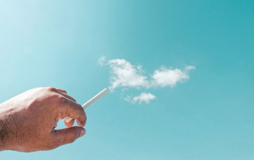 Hand holding a lit cigarette against a blue sky.