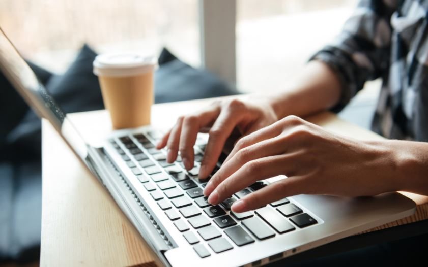 Hands typing on a laptop beside a takeaway coffee cup.