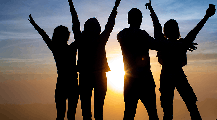 Four people silhouetted against a sunset with their arms raised.